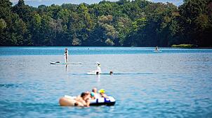 Eine Frau fährt auf einem Stand Up Paddle-Board über den Opfinger See und Badegäste baden im Wasser. In einem Freiburger Badesee soll ...