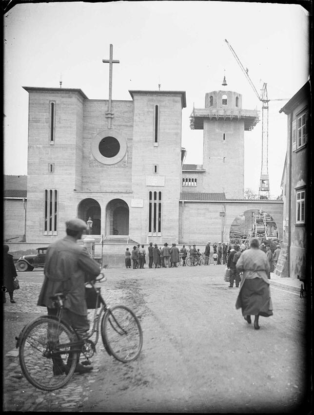 Neugierige beobachten das Ablassen der Kirchenglocke aus dem offenen Kirchturm. Das neue Kirchengebäude steht hier bereits.