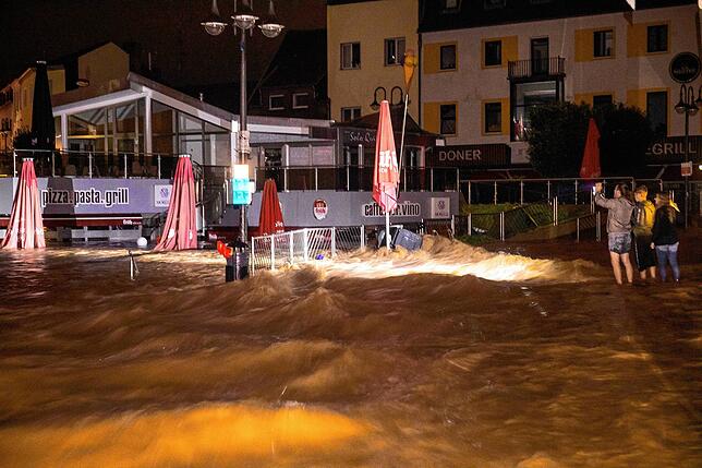 In Euskirchen in Nordrhein-Westfalen beobachten drei Menschen die Wassermassen, die sich über eine Straße und ein Restaurant ausbreiten.