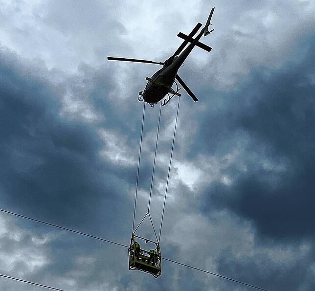Monteure reparieren Seile an Strommästen in einer unter dem Helikopter hängenden Aluminiumgondel.