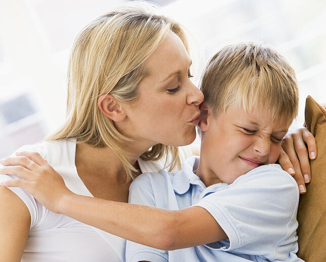 Woman kissing disgusted young boy in living room Küssen verboten: Kinder mögen es nicht immer, wenn Mama einem einen dicken Schmatzer aufdrücken möchte.