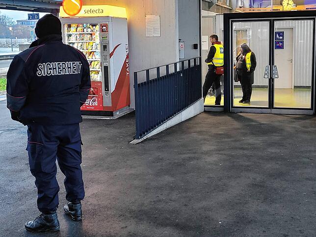 Im Vordergrund ein Sicherheitsmann der SBB. Im Hintergrund die gläserne Vorhalle des Zoll-Gebäudes, in der SBB-Mitarbeiter Bahntickets ...