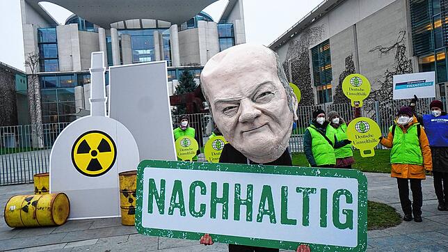 Ein als Bundeskanzler Scholz verkleideter Aktivist steht mit einem Transparent mit der Aufschrift &bdquo;Nachhaltig&ldquo; vor einem ...