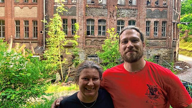 Isabella und Christopher Fuchs vor der Porzellanfabrik, zu der mehrere Gebäude und ein großes Grundstück mit Wald und einer Quelle gehören.