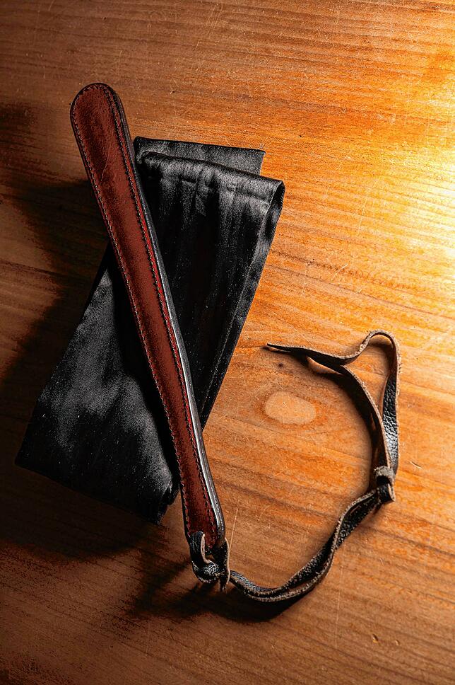 Ein sogenanntes Paddle (zu deutsch: Paddel) liegt auf einer Augenbinde auf dem Holztisch. Mit diesem Paddle versohlen sich Karin, ...