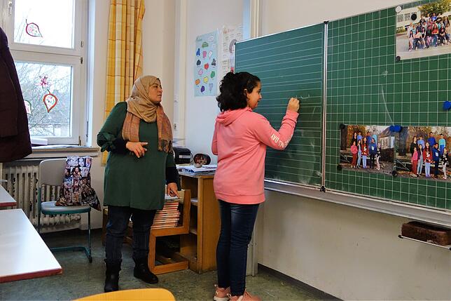 Rahaf schreibt an der Tafel die Buchstaben, die sie als Hausaufgabe geübt hat. Lehrerin Fatema Alali schaut aufmerksam zu.