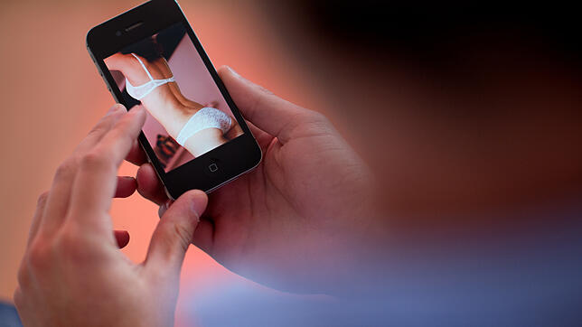 Ein junger Mann hält ein Smartphone, auf dem ein erotisches Foto einer jungen Frau zu sehen ist.