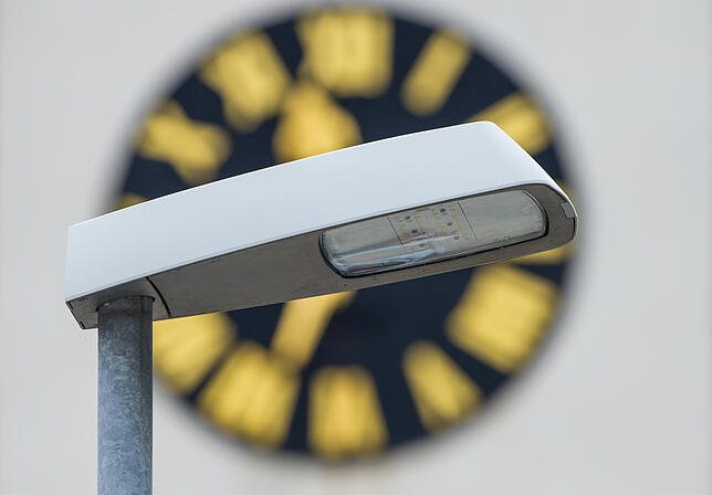 Eine Straßenlampe mit LED-Leuchten steht in Sandhausen (Baden-Württemberg) vor dem Ziffernblatt eines Kirchturms.