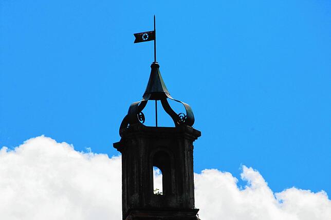 Diese Turmspitze hat eine bemerkenswerte Flagge aus Metall. Sie ist von Weitem bereits zu sehen.