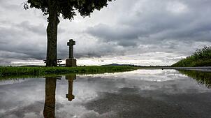 Neben einem Wegkreuz unter einem Baum spiegeln sich dunkle Wolken in einer Regenpfütze.