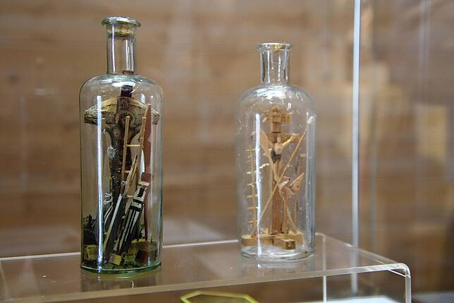 Zwei Igrichte oder Geduldsflaschen stehen in einer Vitrine im Beriech Alltag und Kirche. Die Kreuzigungsdarstellung wurde in mühevoller ...