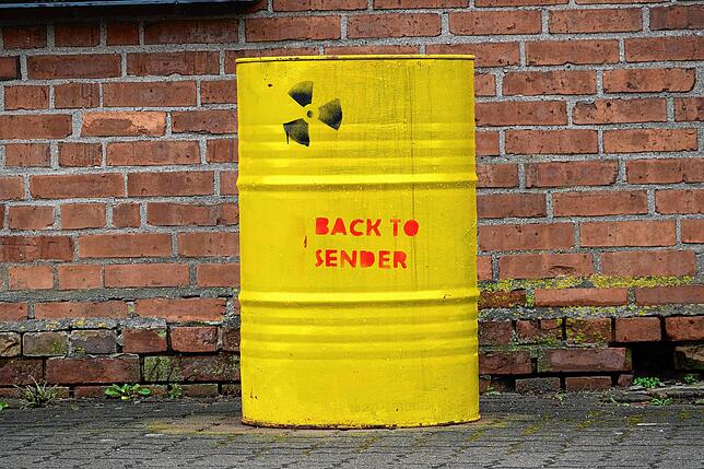 Ein Fass mit der Aufschrift “Back to sender„ und einem Atomzeichen steht vor einem Haus.