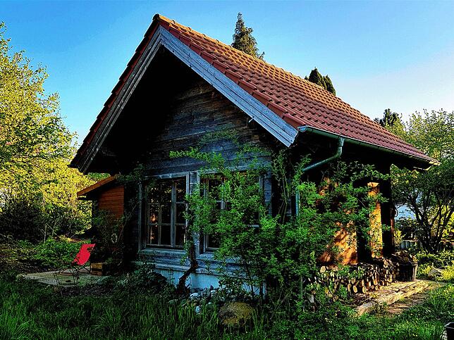 Hintenraus hat das Häuschen große Fenster – raus in den Garten. Holz lagert an der rechten Wand und auf der anderen Seite im Schuppen.