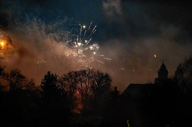 Das Feuerwerk leuchtet durch die Bäume hindurch. Deren Äste und die Gebäude werden aus der Finsternis herausgeschält wie Schattenrisse.