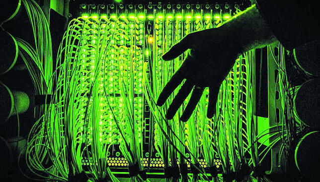 Schnelles Internet läuft über Glasfaser: Das Bild zeigt eine Hand vor einen Verteilerpunkt, in dem zahlreiche Glasfaserkabel ...