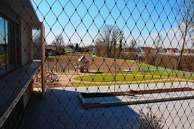 Ausblick vom Fluchtweg durch das Edelstahlnetz auf den Schulhof und Teile des Spielplatzes, hinten im Bild die Mühle, rechts die Rotach.