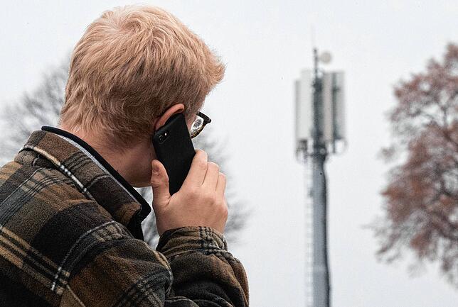 Ein Mann telefoniert mit einem Smartphone neben einer Basisstation (Mobilfunkmast). Oft sind die Masten Gegenstand von Diskussionen.