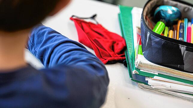 Eine Mund-Nasen-Bedeckung liegt während einer Unterrichtsstunde neben einem Mäppchen und Schulbüchern auf einem Schultisch. Auch auf dem ...