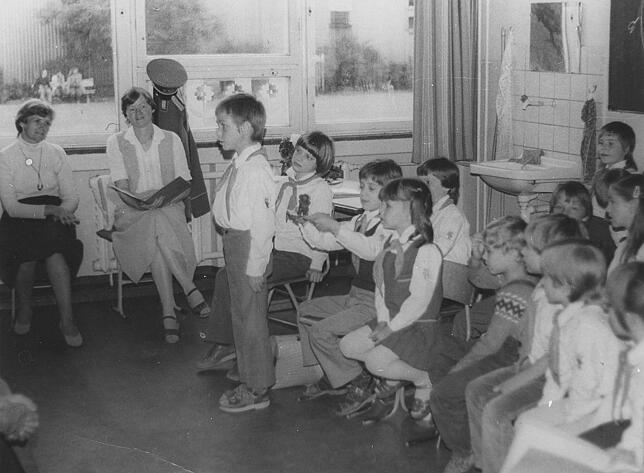 Jungpionier Jörg (stehend) trägt ein Gedicht vor, seine Mitschüler und Erzieher hören zu.