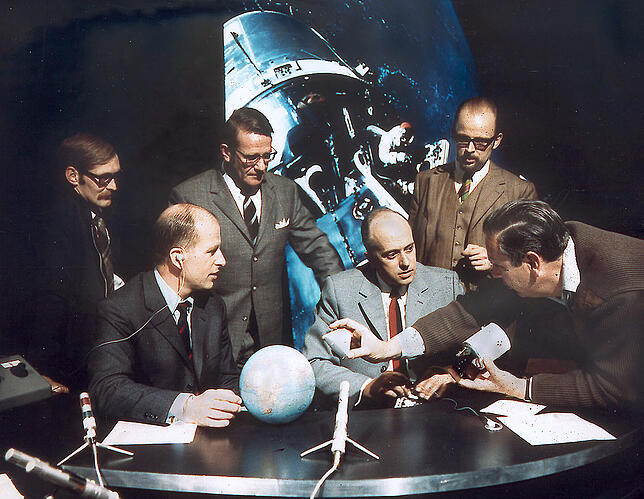 Regiebesprechung im Appollo-Sonderstudio. Vorne ein Globus, im Hintergrund ein Foto des Raumschiffs von Apollo 9.