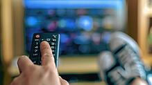 Eine Frau ändert beim Fernsehschauen den Sender mit der Fernbedienung. Am 1. Juli fällt das Nebenkostenprivileg von Kabel-TV.