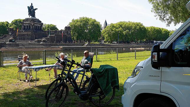 Touristen sitzen auf einem Campingplatz in Koblenz vor ihren Wohnmobilen in der Sonne.