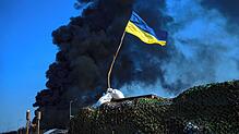 Eine ukrainische Flagge weht Ende März auf einem Kontrollpunkt, während schwarzer Rauch aus einem Treibstofflager der ukrainischen Armee ...