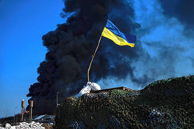Eine ukrainische Flagge weht Ende März auf einem Kontrollpunkt, während schwarzer Rauch aus einem Treibstofflager der ukrainischen Armee ...