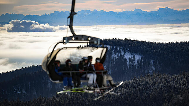Wintersportler sitzen vor der Alpenkulisse am Feldberg in einem Sesselllift.