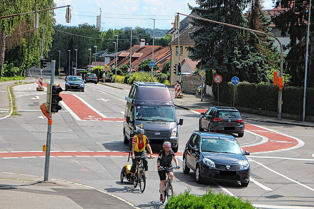 Autofahrer überholen Radler. Der Fahrer des vorderen Autos nutzt die Linksabbiegespur des Gegenverkehrs.