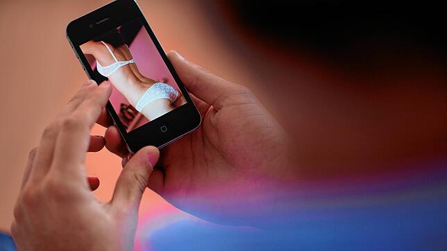 Das Symbolfoto zeigt einen jungen Mann, der auf ein Smartphone schaut, auf dem ein erotisches Foto einer jungen erwachsenen Frau zu ...