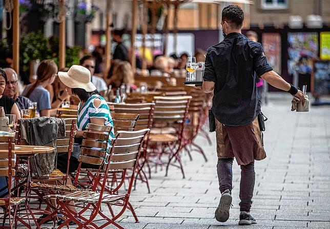 Eine Servicekraft läuft im Außenbereich eines Restaurants an Gästen vorbei. Der Fachkräftemangel nimmt drastische Formen an
