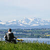 09.05.2021, Baden-Württemberg, Meersburg: Eine Frau sitzt in ihrem Campingstuhl an einem Parkplatz, während im Hintergrund die Alpen in ...