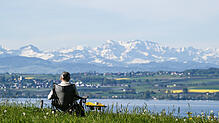 09.05.2021, Baden-Württemberg, Meersburg: Eine Frau sitzt in ihrem Campingstuhl an einem Parkplatz, während im Hintergrund die Alpen in ...