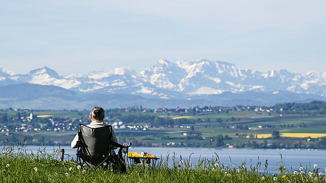 09.05.2021, Baden-Württemberg, Meersburg: Eine Frau sitzt in ihrem Campingstuhl an einem Parkplatz, während im Hintergrund die Alpen in ...