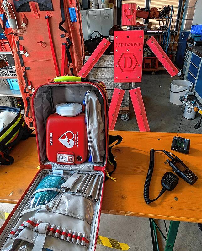 Vorne ist ein Rucksack mit Defibrillator geöffnet und daneben liegen neue Funkgeräte. Unten steht die Übungspuppe für das Sonar.