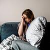 Eine junge Frau mit Long-Covid-Symptomen wie Erschöpfung und Kopfschmerzen sitzt auf Hause auf dem Sofa.