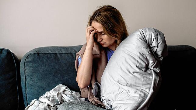 Eine junge Frau mit Long-Covid-Symptomen wie Erschöpfung und Kopfschmerzen sitzt auf Hause auf dem Sofa.