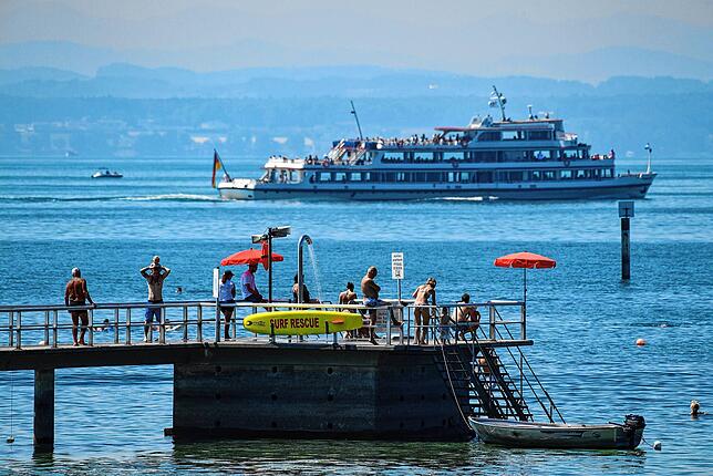 Mehrere Badegäste laufen im Strandbad in Friedrichshafen auf den Steg zum Einstieg in den Bodensee. Der See ist Touristenmagnet aber ...