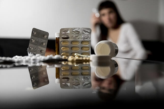 Die gestellte Szene zeigt Tabletten auf einem Glastisch, während eine Frau dahinter sitzt. Schmerzmittel haben ihre Licht- und ...