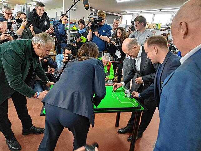 Action vor laufenden Kameras beim Tischfußball. Kanzler Scholz, rechts am Tisch, spielt den Ball.