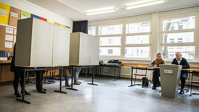 Ehrenamtliche Wahlhelfer sitzen in einem Wahllokal vor einer Urne, während in den Wahlkabinen gewählt wird.