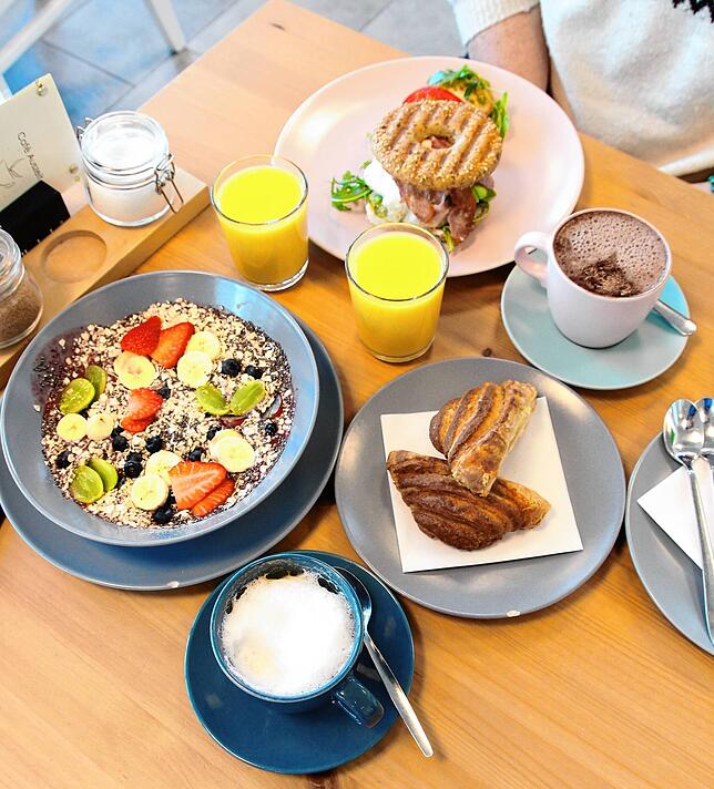 Auf dem Tisch stehen ein Smoothie Bowl Frühstück (Cappuccino, Orangensaft und Bowl), ein Hamburger Frühstück (Heiße Schokolade, ...