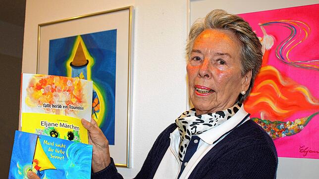 Die Künstlerin Liane Hoyer zeigt drei ihrer Märchenbücher, die ihre Bilder und Texte enthalten. Im Hintergrund hängen zwei ihrer Bilder. ...