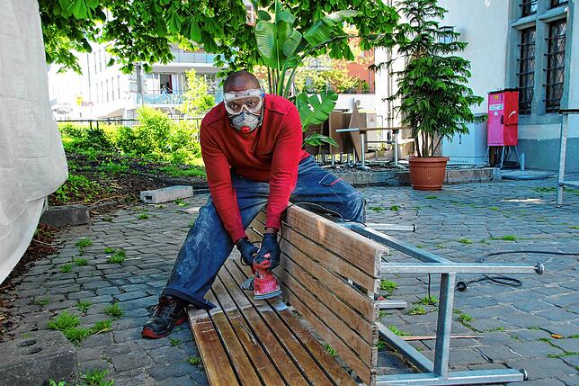 Der Auszubildende Augustin Nwachukwu schleift, streicht und lackiert sämtliche Bänke, Stühle und Tische der Konzil-Terrasse. Eine ...