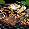 Spareribs, Gemüse, gefüllte Hühnerbrust und Garnelen liegen auf einem Grill. Gewendet wird gerade ein Soja-Steak.