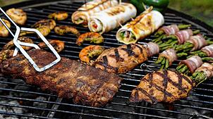 Spareribs, Gemüse, gefüllte Hühnerbrust und Garnelen liegen auf einem Grill. Gewendet wird gerade ein Soja-Steak.