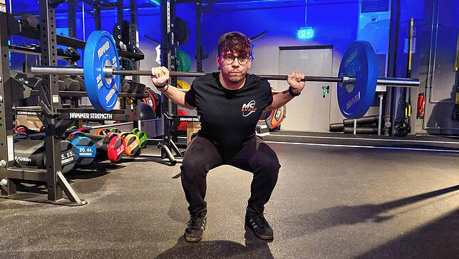 Trainer Oliver Leyva Puig (22) zeigt Kniebeugen mit einer Langhantel. Diese Übung kräftigt Oberschenkel, Gesäß, Waden, Rumpf und Schultern.
