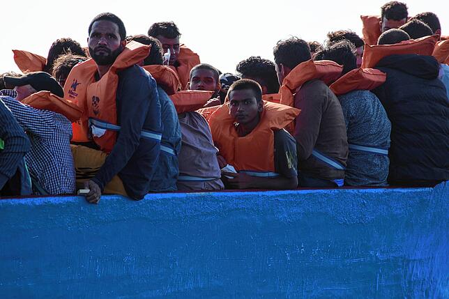 Zusammengepfercht sitzen die Geflüchteten eng beieinander auf ihren Holzbooten. Die Boote drohen zu kentern, weil sie hoffnungslos ...